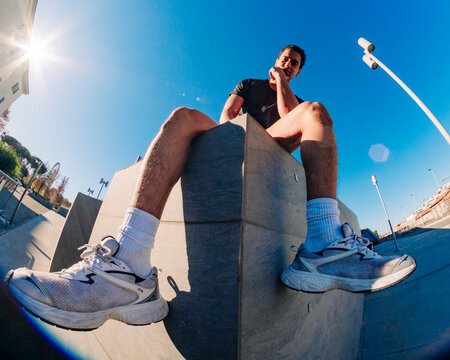 A low-angle fisheye view of a young man in athletic attire seated on a concrete ledge in an urban environment with clear skies and bright sunlight in the background.