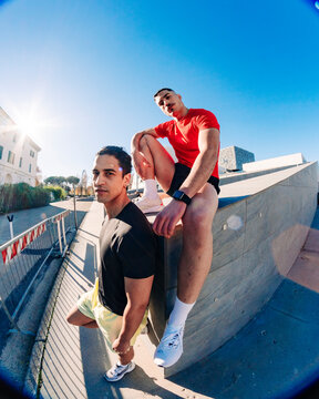 Two young men, one sitting on a ledge in a red shirt and the other in yellow shorts, in an outdoor urban space under a clear blue sky with a fisheye perspective capturing the setting.