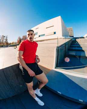 Male runner in a bright red shirt leans casually against a modern architectural structure under a clear blue sky on a sunny day, exuding confidence and style in a contemporary setting.