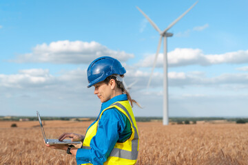 Female Engineer works on laptop in wind farm during daylight hours surrounded by golden fields