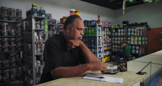 Male shop owner using calculator at counter, focused on finances and business management, small retail entrepreneur balancing expenses and planning