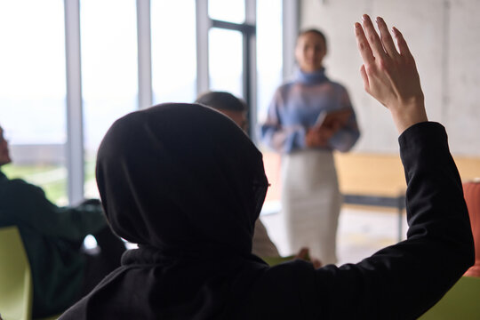 A Muslim businesswoman, dressed in professional attire and wearing a hijab, raises her hand during a business meeting to ask a question to the educator leading the session