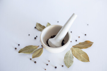 White mortar with black pepper and bay leaves on a white background.