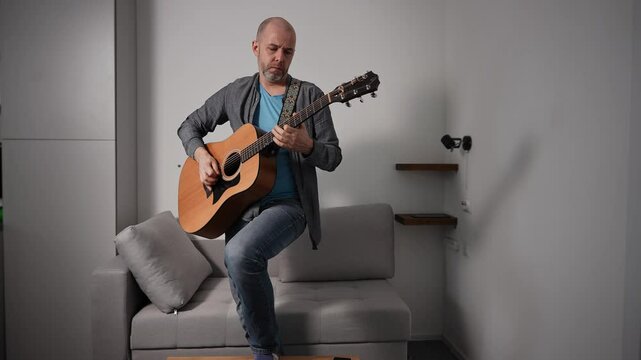 A man passionately plays an acoustic guitar while standing in a simple room. His facial expression reflects the emotional intensity of his music, with a strap securing the guitar around his shoulder.