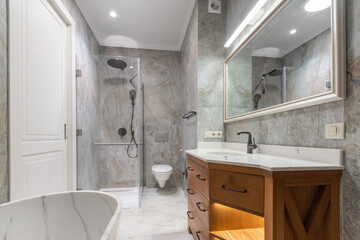 Elegant design of the bathroom, decorated with marble tiles. Wooden cabinet with sink, .illuminated mirror. Glass shower cabin.