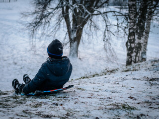 A small child in winter clothing slides down a hill on a blue sled in a snowy park surrounded by...