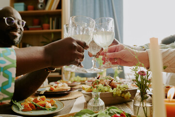 Medium close up of glasses with champagne in hands of biracial family members above festive table with gourmet dishes