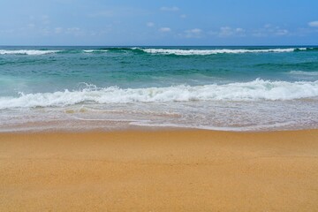 The image depicts a sandy beach with turquoise ocean waves under a blue sky. The shoreline features light brown wet sand, with gentle, small white waves lapping at the beach.