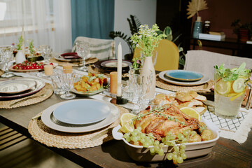 Medium close up of table with ceramic bowl with roasted chicken with juicy oranges and grapes, different types of salad and kitchenware on table