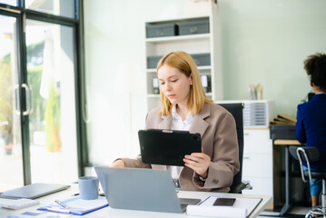 Obraz premium Young woman, smiling and working at her desk in an office environment. She is wearing a business suit