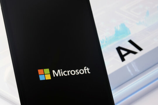 Microsoft logo is displayed on the smartphone, while a futuristic background  with AI ( Artificial Intelligence ) sign is shown in the background on the computer screen.
