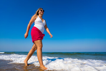 Beautiful middle-aged woman walking on sandy beach in summertime. Physical activity on beach. Side view