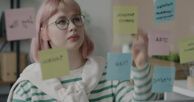 Pensive young woman working with sticky notes thinking reading memos on glass board in office. Businessperson and workplace concept.