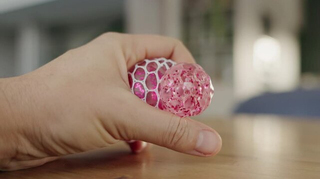 A close-up of a hand pressing a pink squishy stress ball covered in a white mesh, placed on a wooden tabletop.