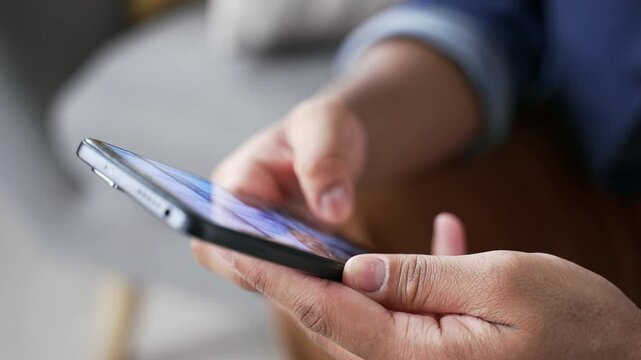 Close up of male hands holding mobile phone sitting on sofa at home. Man uses smartphone, browses social media, scrolls searches for information on the internet, online web user of technology