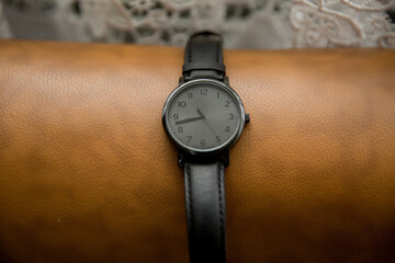 A black watch with a black band sits on a brown surface. The watch is set to the time of 10:00