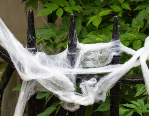 Artificial spider webs draped over a black iron fence with pointed tips, surrounded by green...