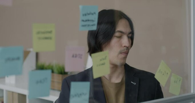 Asian businessman working with information on sticky notes reading looking at glass board in office. Workplace and businesspeople concept.