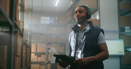 African American Logistics Coordinator in Headset Chats with Customer, Uses Tablet Computer, Looks...
