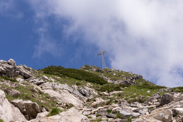 Fototapeta premium A part of the mountain slope with bushes and stones, and a road leading to the top of the mountain, where there is a large cross against the sky with white clouds.