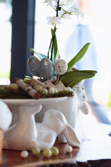A tall vase arrangement with green leaves, white orchids, and speckled decorative eggs, creating a sophisticated Easter-themed centerpiece with natural elegance in soft natural light