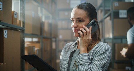 Female Logistics Coordinator with Tablet Computer Chats with Customer on Call Using Mobile Phone in Warehouse Facility. Sorting Center Employee Working with Cardboard Boxes in the Background. Close Up