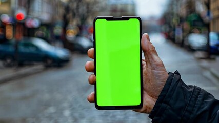 A hand displays a smartphone with a green screen while standing in a deserted urban street. The background features blurred cars and overcast skies, giving a modern feel.