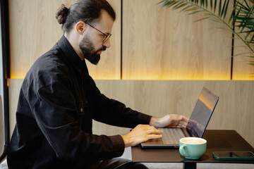 A stylish bearded man with glasses and a laptop sits at a table in a coffee shop and works remotely