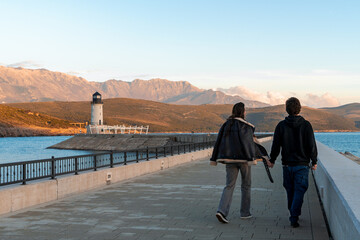 Fototapeta premium A couple walks hand in hand along a scenic coastal promenade, heading towards a lighthouse at sunset. The picturesque view features calm blue waters, rugged hills, and a warm golden light