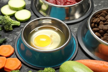 Natural and dry pet food in feeding bowls among products on dark textured table, closeup