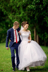 A bride and groom are walking together in a park. The bride is wearing a white dress and the groom is wearing a blue suit