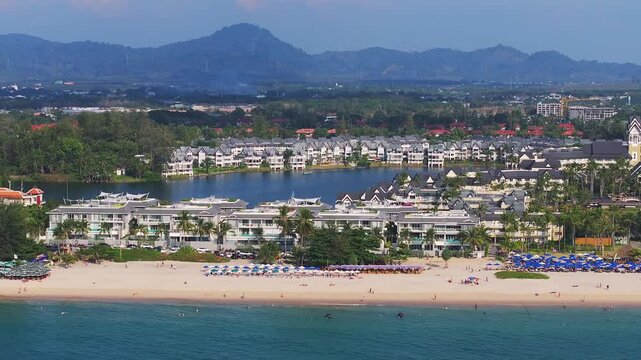 Aerial view of Phuket Island, Thailand, showing a vibrant beach, blue umbrellas, a resort area with white buildings, and a red roofed landmark.