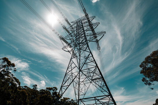 High voltage power lines stretch to the sky under a bright sun with swirling clouds.