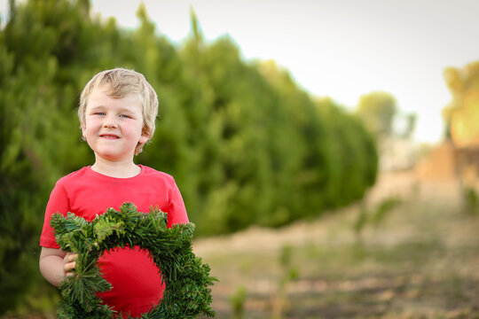 Little boy standing in front of row of Christmas trees on farm holding wreath