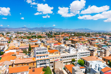 Fototapeta premium Aerial view of Nicosia capital city of Cyprus, featuring a mix of modern and traditional buildings with tiled roofs. Cityscape is set against a backdrop of distant mountains and scattered clouds sky