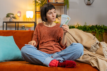Smiling young woman counting money dollar banknotes using smartphone calculator app on sofa in room...