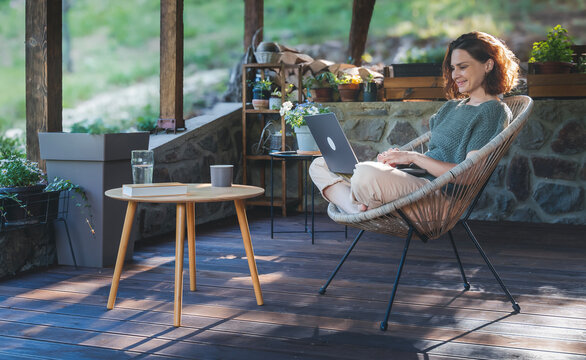 Young adult Caucasian woman sitting on cottage terrace using laptop in summertime, online work