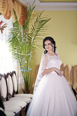 Fototapeta premium A woman in a white wedding dress stands in front of a potted plant. She is wearing a veil and is posing for a picture. The room has a yellow wall and a few chairs