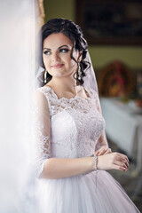A woman in a white wedding dress is standing in front of a wall. She is smiling and looking at the camera