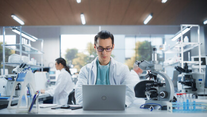 Genetic Research Scientist Working on a Laptop Computer in an Applied Science Laboratory. Portrait of a Handsome Young Multiethnic Lab Engineer in White Coat at Work