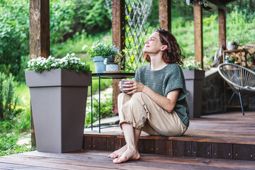 Young happy smiling Caucasian woman sitting on the wooden terrace of a country house enjoying the...