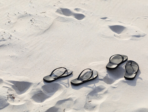 two pairs of black thongs on white beach sand with footprints