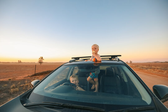 Little kids hanging out in car with one sticking out sunroof eating an apple on a country road