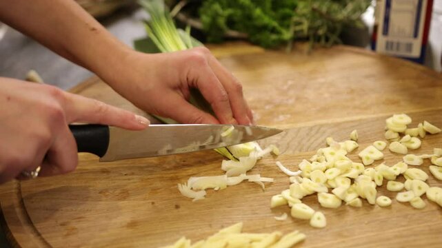 Fresh vegetables, Leek preparation, Knife slicing. Image captures hands using knife to chop leek on wooden cutting board, with garlic and fresh veggies nearby.