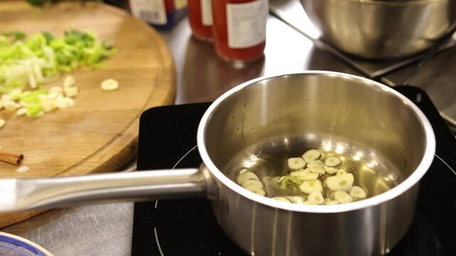 Sliced garlic, Stainless pan, Cooking garlic. Sliced garlic cooking in stainless steel pan on stove, with chopped vegetables on wooden cutting board nearby.