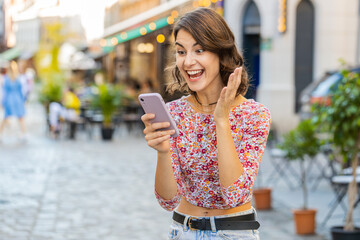 Fototapeta premium Young woman use mobile smartphone celebrating win good message news, lottery jackpot victory, giveaway online, play game outdoors. Happy girl tourist standing in urban city street. Town lifestyles