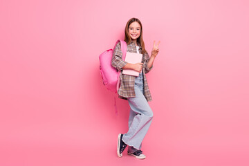 Charming young schoolgirl posing with a pink backpack and notebook against a pink background,...