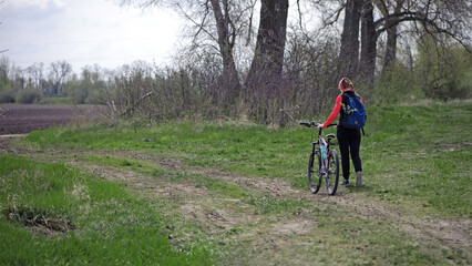 Fototapeta premium a girl with a bicycle walks a field trail. Woman riding a mountain bike in the forest. Hiker woman with backpack. one young woman riding a bike in nature. rolling the bike in his hands. back view.