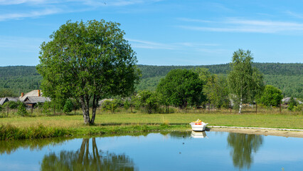 Small non-motorized or motorless white pleasure boat on shore of pond or lake in village or rural area. Rural pastoral landscape. Outdoor recreation in good environment