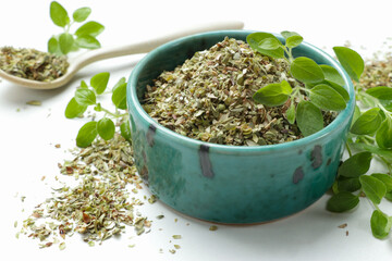 Dried oregano in bowl, spoon and green leaves on white table, closeup
